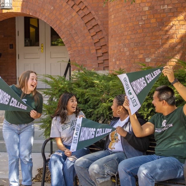 A group of four MSU Honors College students in front of the red brick entrance to Eustace-Cole Hall. Three students are sitting on a black bench, while another is standing. The students are in jeans and various MSU t-shirts, holding green and white Honors College pennants.