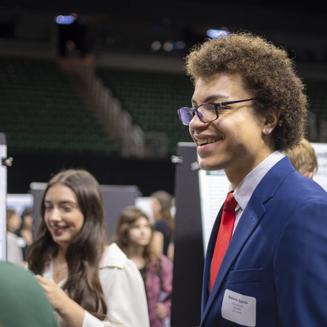 Image of Marcos Garcia standing and smiling in a navy blazer, collared shirt, and red necktie at the Breslin Center on MSU’s campus for the 2026 University Undergraduate Research and Arts Forum. Research posters and other people are in the background.