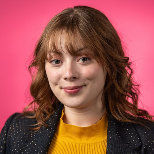 Michigan State University Honors College student and Rhodes Scholarship nominee Anastasia Breen softly smiling in a yellow shirt and black dotted blazer, against a hot pink background.