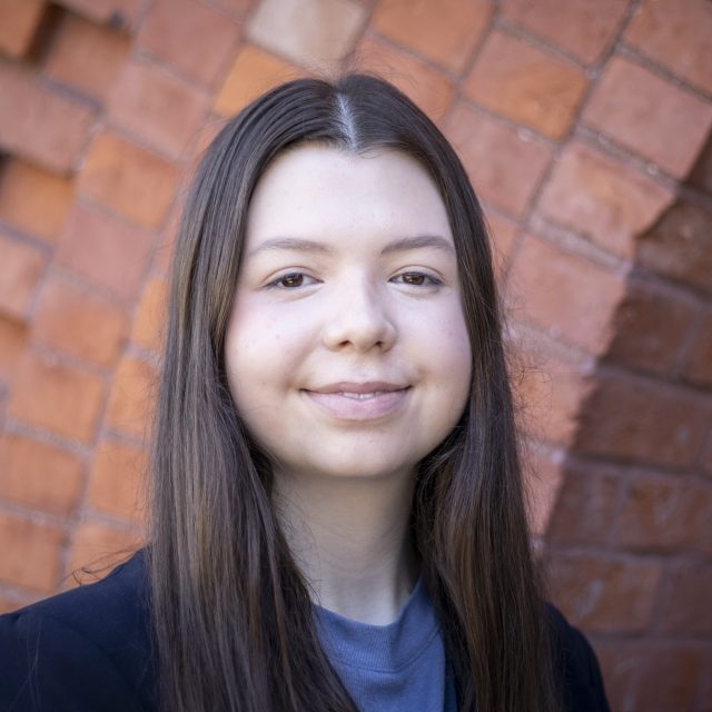 MSU Honors College student and Rhodes Scholarship nominee Eleanor Pugh wearing a black blazer, softly smiling and standing in front of the red brick entrance to Eustace-Cole Hall.