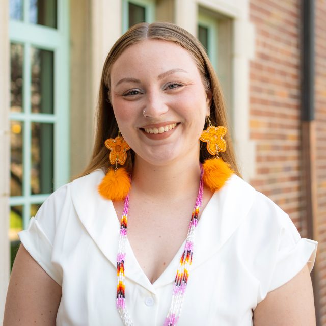 MSU Honors College alumna and Rhodes Scholarship nominee Kaylin Casper wearing orange earrings, a colorful beaded necklace, and a white top. Casper is standing and smiling in front of Campbell Hall.